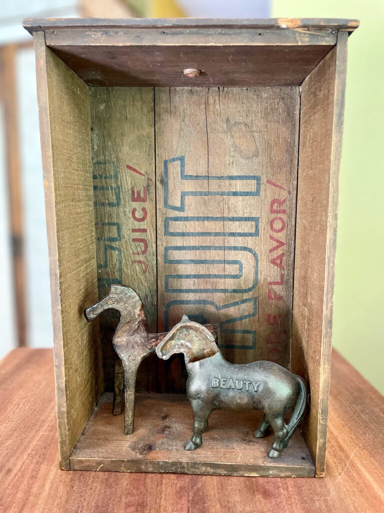 Sculpture: Small wooden box standing on end. Small 2-D horses standing inside it.