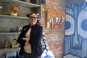 Woman with glasses wearing a black jacket stand smiling in front of a folk art display.