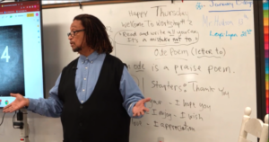 Photo of man standing in front of a white board, wearing a blue shirt and black sweater vest.