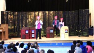 Actors on a stage costumed as the Three Little Pigs perform among their three houses.