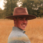 Smiling young white man from shoulders up. He wears a brown cowboy hat and a blue collared shirt, and he's in a wheat field.