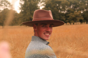 Smiling young white man from shoulders up. He wears a brown cowboy hat and a blue collared shirt, and he's in a wheat field.