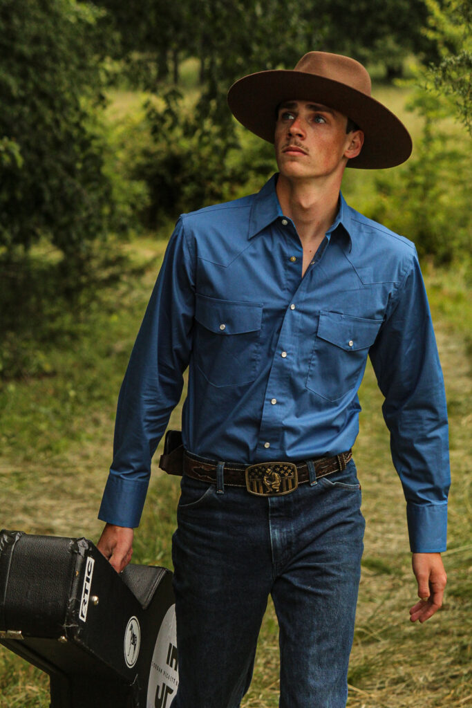 Young white man in a cowboy hat, jeans, and a blue button-down shirt. He's outside and looks seriously to his right as he walks.