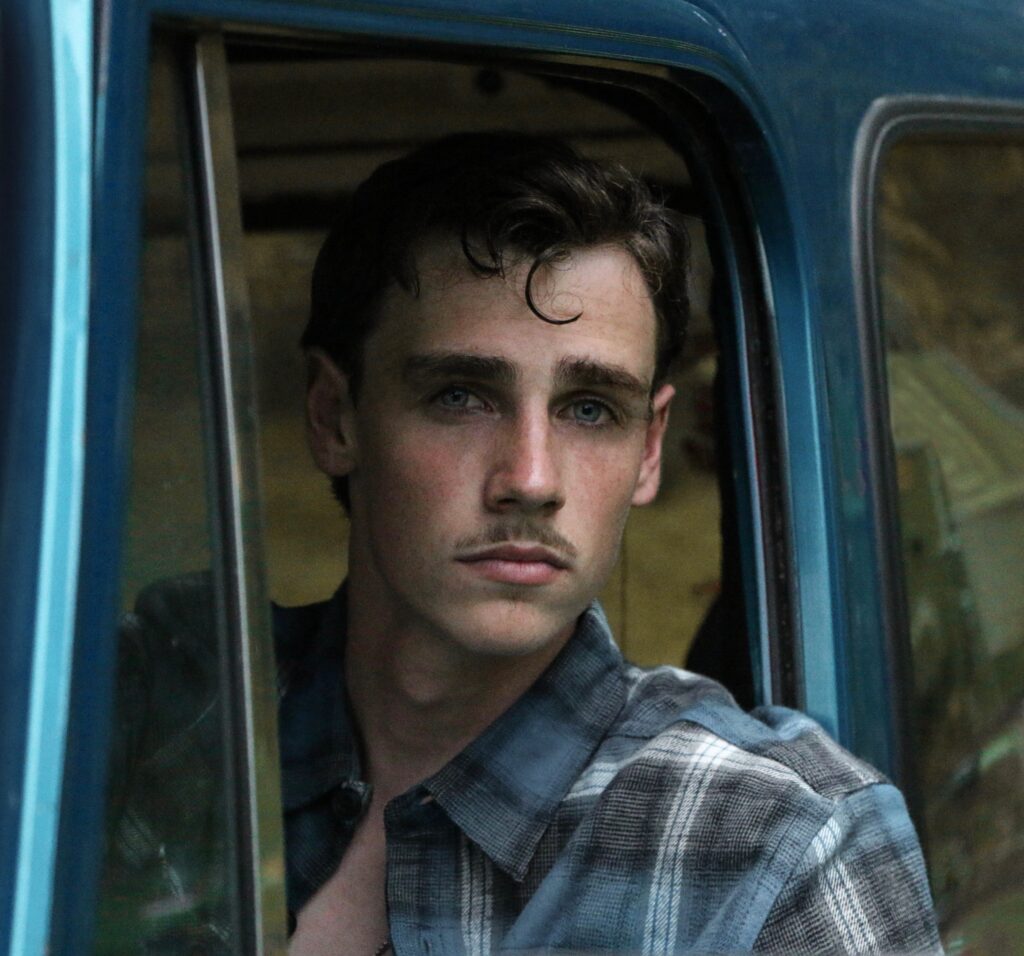 Young white man with a mustache looking out the driver's side window of a blue truck. He's got brown hair, and he wears a plaid shirt and serious expression.