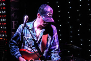 Young white man in a ball cap and plaid shirt, looking down at his guitar as he performs onstage.