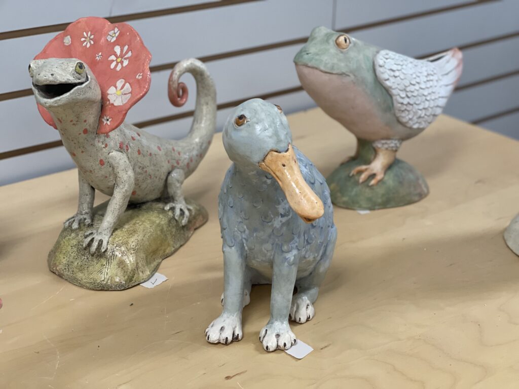 Three sculptures on a table: a frog creature with wings, a bearded lizard, and a duck-dog