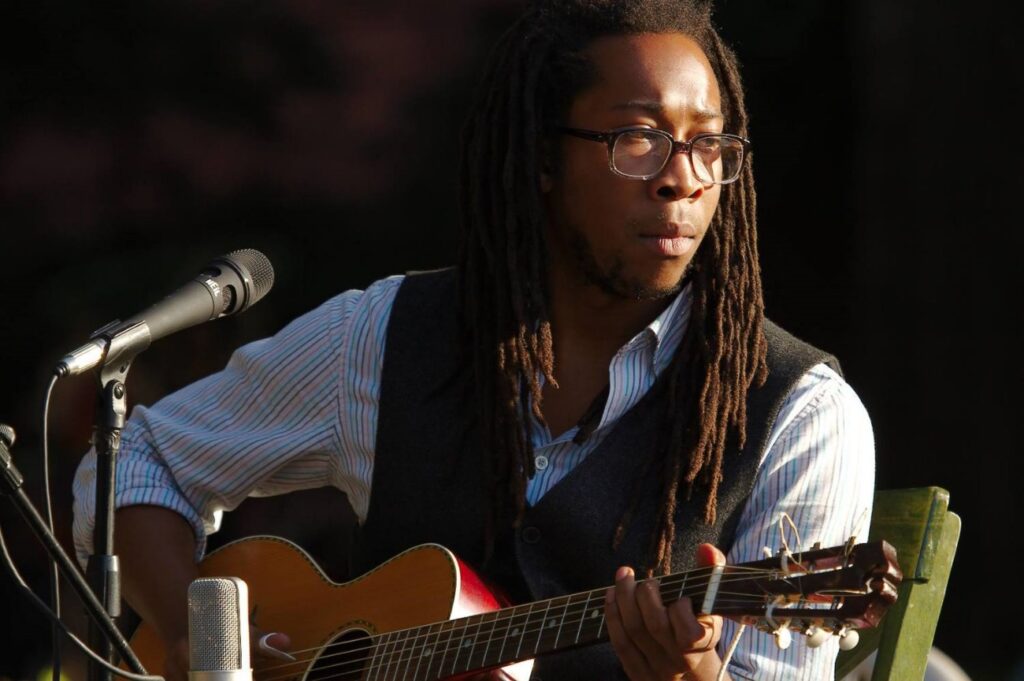 A black man with locs and glasses wearing a white button down and black vest playing guitar