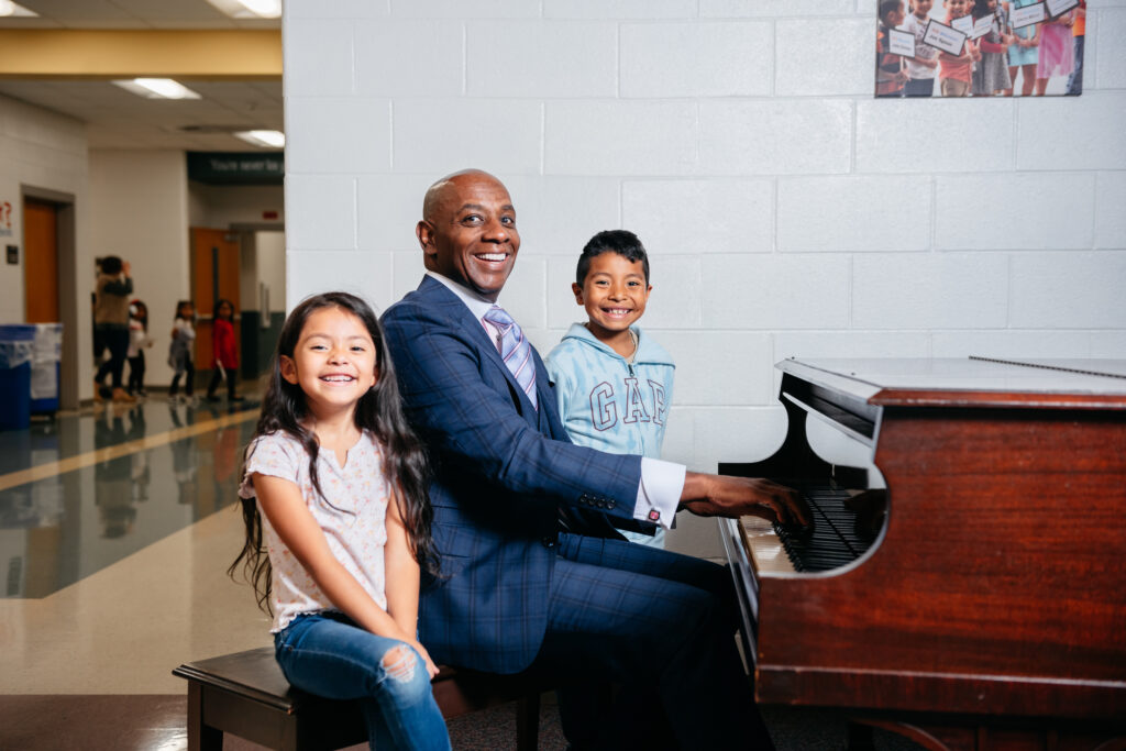A black man in a blue suit is seated at piano bench with two elementary school kids, a girl to the left and a boy to the right. He is playing piano and all are smiling.