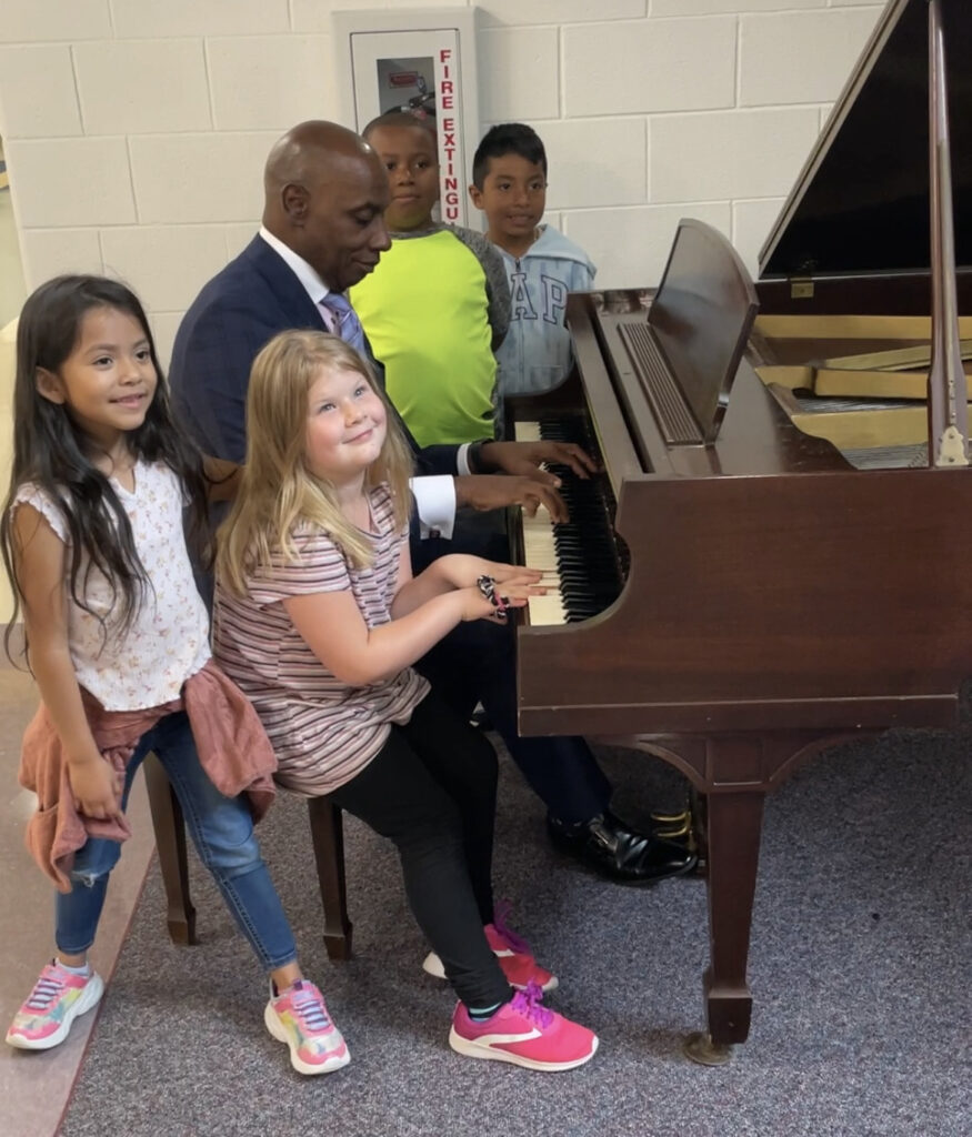 A black man in a blue suit plays piano in the center of a group of elementary school children. From left to right: a girl with long dark hair in jeans and a white shirt, a girl with long blonde hair sitting and playing along, a boy in bright yellow, and a boy in a Gap hoodie.