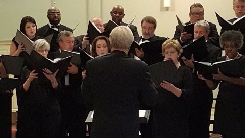 An ensemble of performers wearing uniform black attire are holding choral notebooks and singing. In the foreground, a conductor with short white hair leads the group.