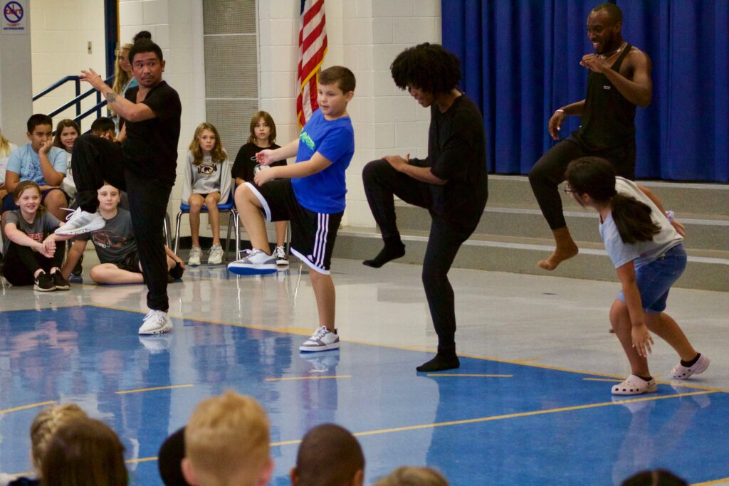 Professional dancers perform in front of an audience of children inside a school's multipurpose room.