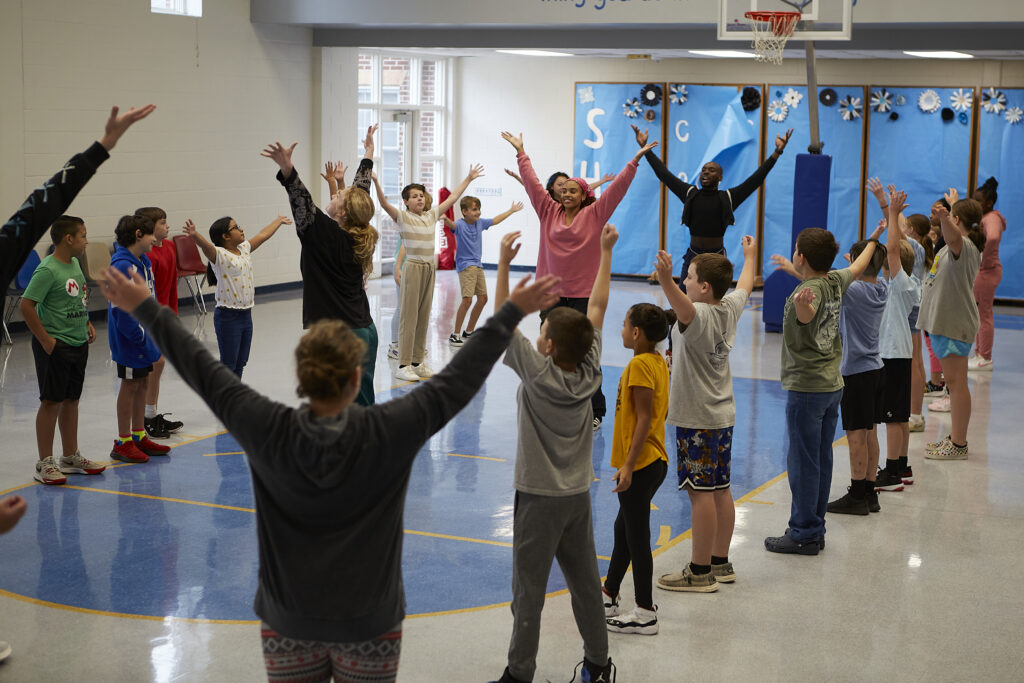 Children stand with their hands raised upwards alongside instruction from professional dancers inside a school multi-purpose room. 