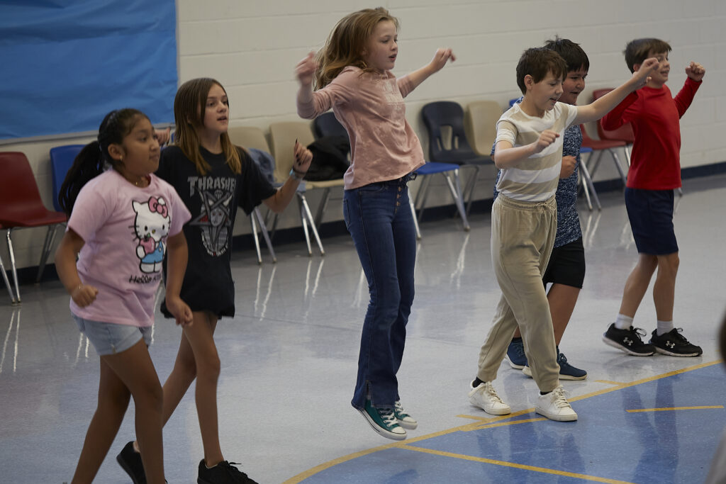 Students play a game of making different shapes with their bodies through dance with the help of professional instructors inside a school multipurpose room.