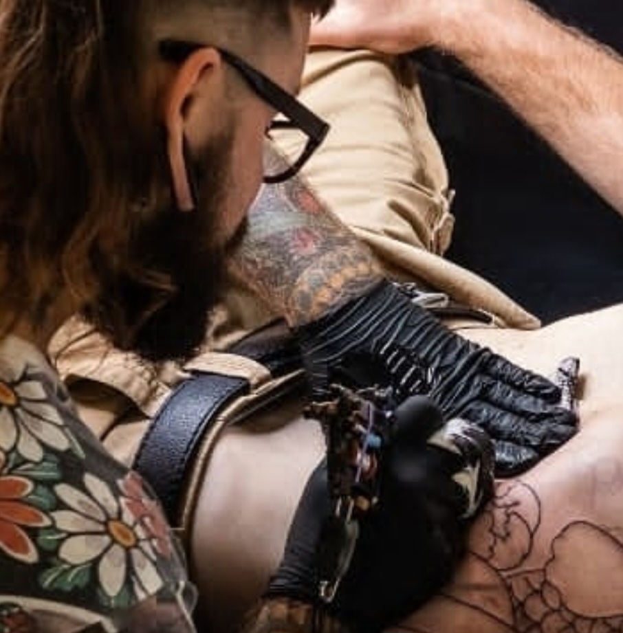 A bearded man with glasses is tattooing someone's back