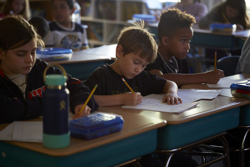 Phillip Shabazz, a tall Black man, instructs fifth grade students on poetry writing in a dimly lit and cozy classroom