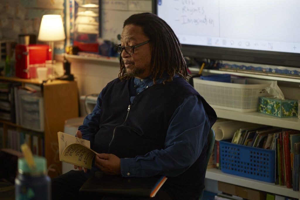 Phillip Shabazz, a tall Black man, instructs fifth grade students on poetry writing in a dimly lit and cozy classroom