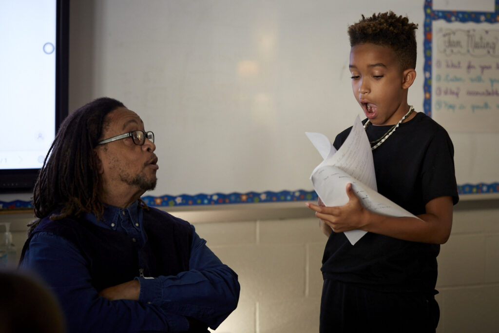 Phillip Shabazz, a tall Black man, instructs fifth grade students on poetry writing in a dimly lit and cozy classroom