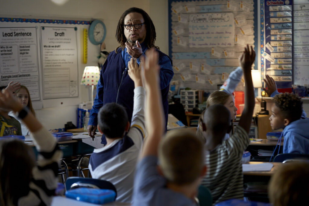 Phillip Shabazz, a tall Black man, instructs fifth grade students on poetry writing in a dimly lit and cozy classroom