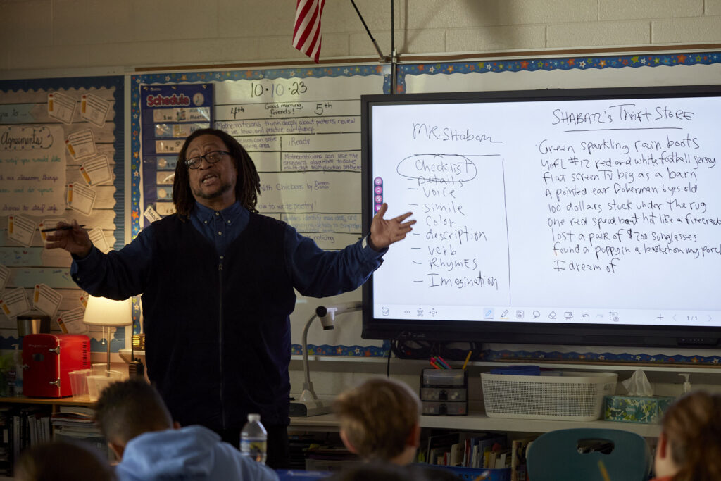 Phillip Shabazz, a tall Black man, instructs fifth grade students on poetry writing in a dimly lit and cozy classroom