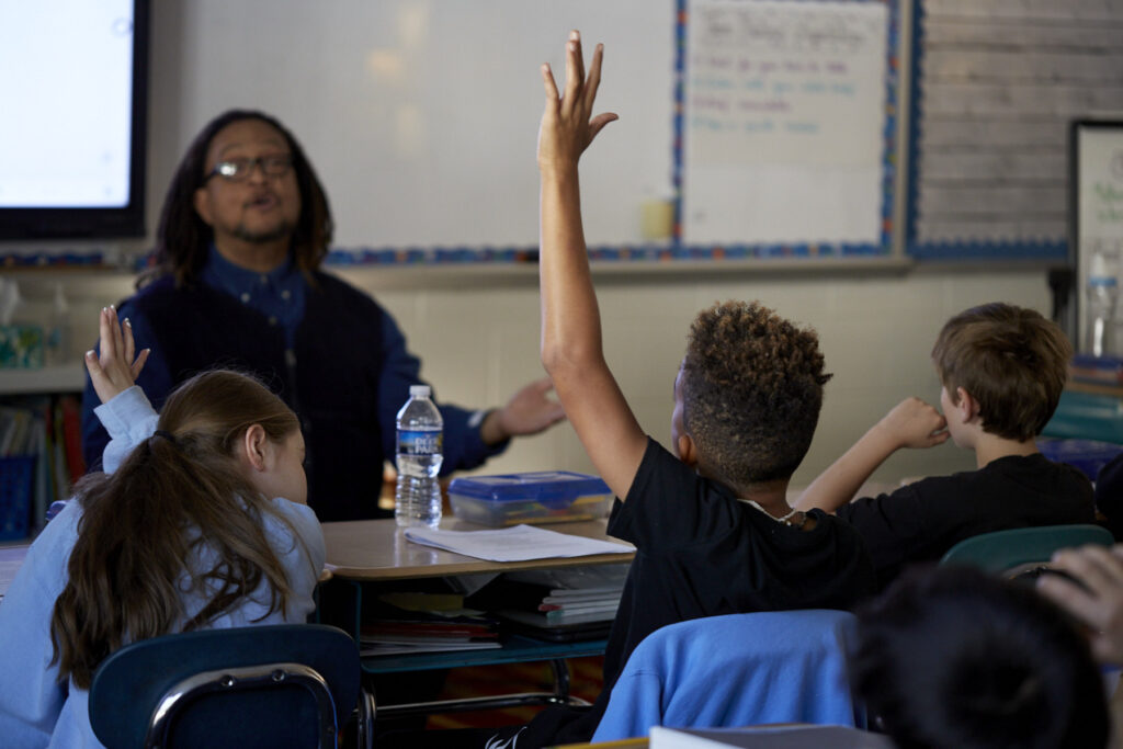 Phillip Shabazz, a tall Black man, instructs fifth grade students on poetry writing in a dimly lit and cozy classroom