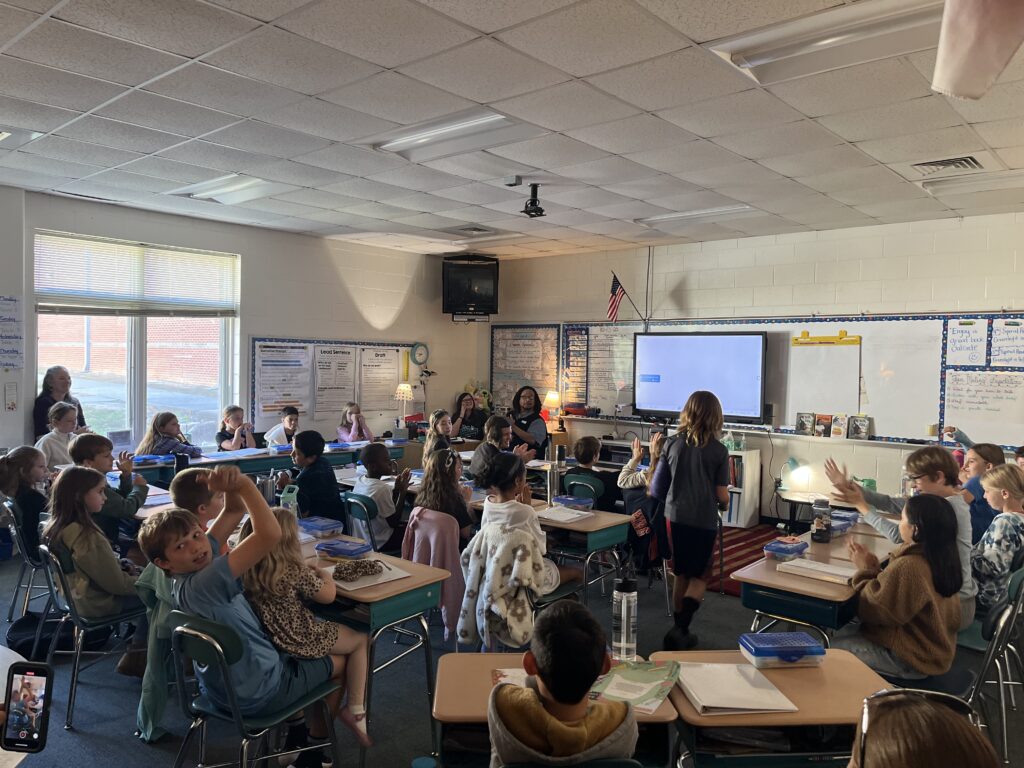 Phillip Shabazz, a tall Black man, instructs fifth grade students on poetry writing in a dimly lit and cozy classroom