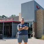 a white woman with a casual mohawk, blue t-shirt, and light blue shorts stand with tattooed arms crossed outside of a restaurant