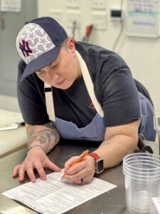 A chef in a Yankees hat, blue apron and tattoos, leaning over a table planning a menu