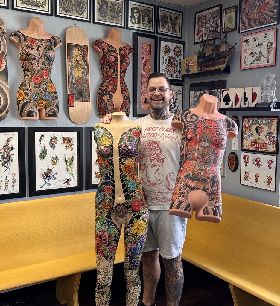 A man with tattoos and glasses displays two of his Japanese body suits in front of a gallery of tattoo art.
