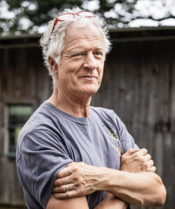 an older man with short gray hair an asymmetrical smile is wearing a heather gray tee shirt and crossing his arms