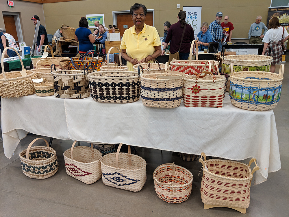 an older black woman standing behind a display table full of multicolored baskets at a convention.