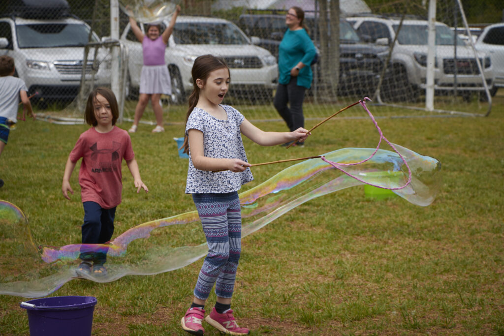 Girl holding sticks with string attached to create giant bubble while boy watches chases it from behind