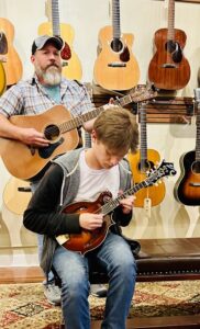 two musicians playing music in front of a wall of guitars, a bearded man in a plaid shirt stands playing guitar and a young man is seated in front playing mandolin.