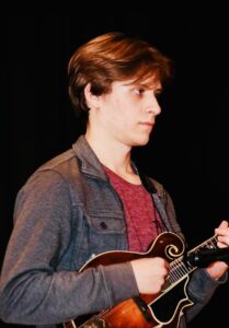 a young man with a red shirt and charcoal button down stands in profile playing the mandolin.
