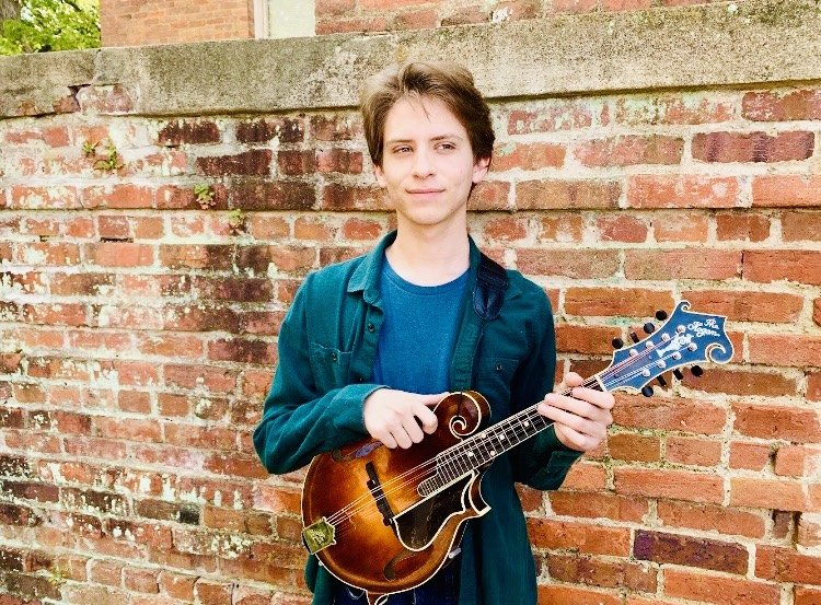 a young man standing in front of a brick wall wearing a blue collared shirt and holding a mandolin