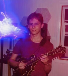 a young man stands holding a mandolin. a flash of light appears in blue and purple before him.