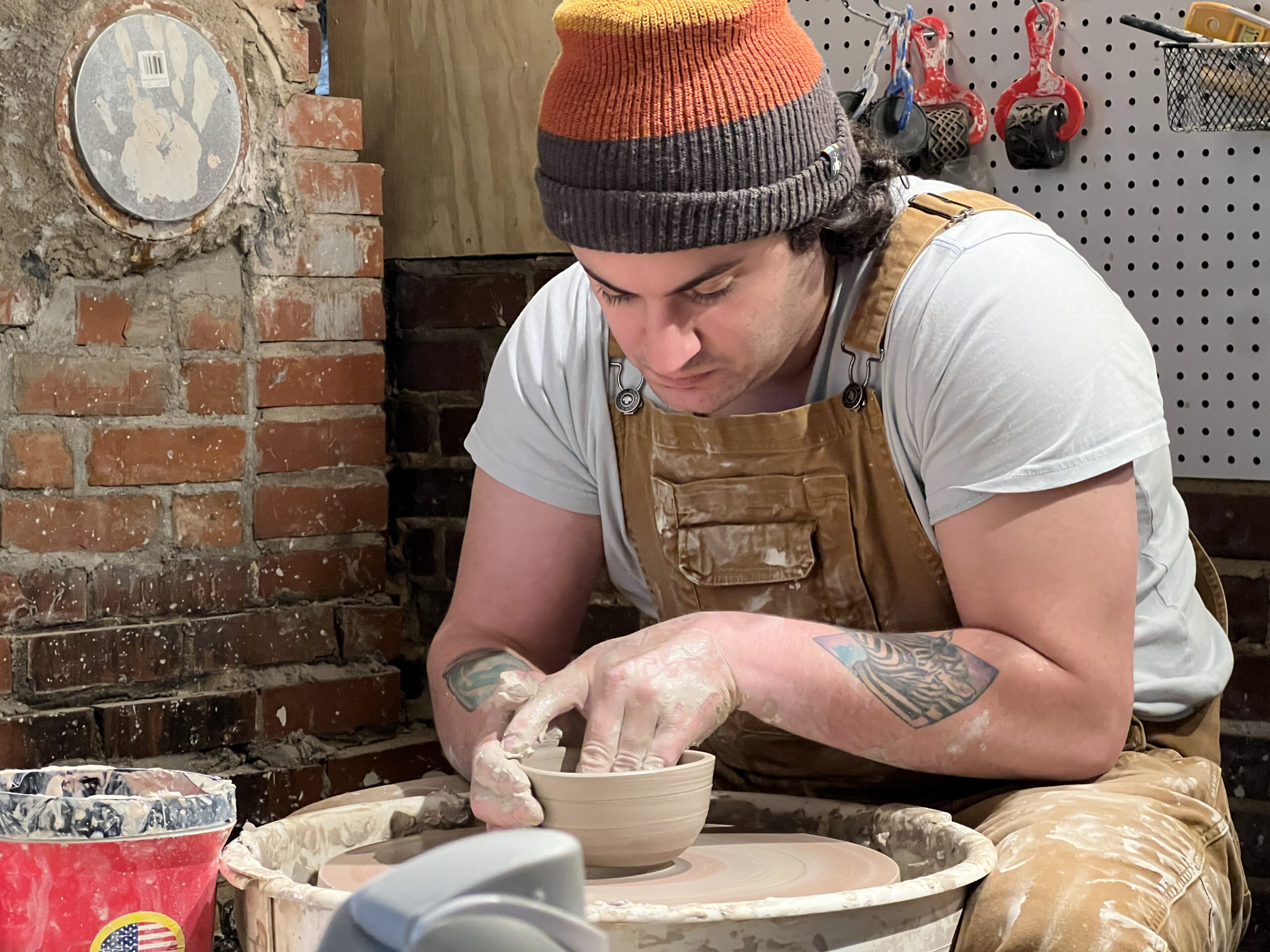 an artist wearing a pale tee shirt, paint spattered overalls, and an orange beanie is working over a potter's wheel. 