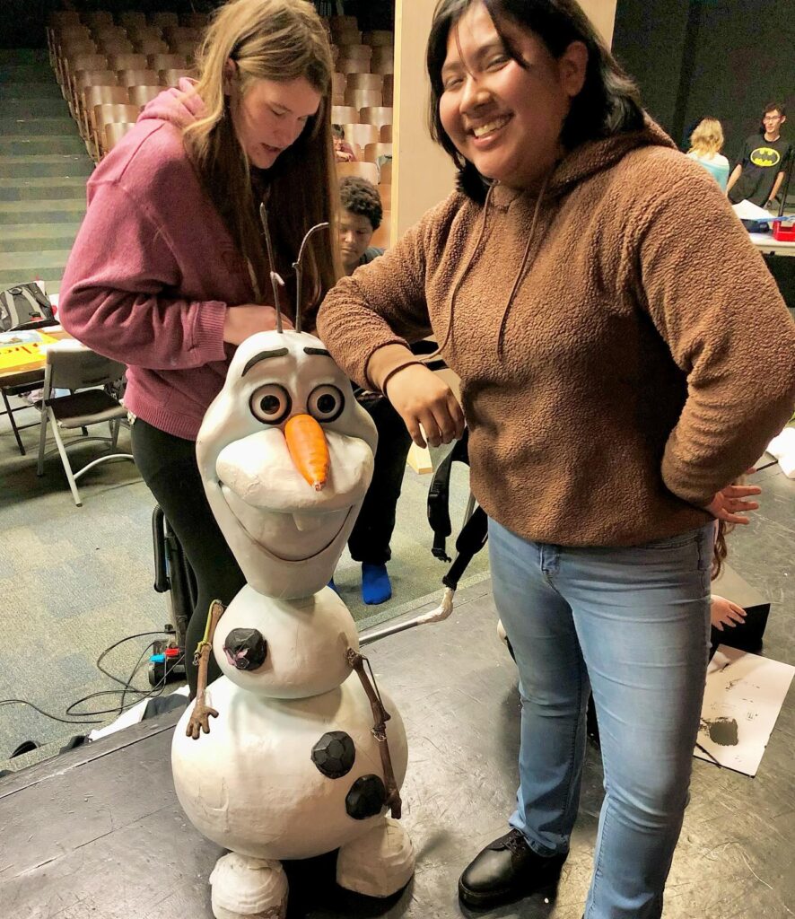 two smiling teenage girls in hoodies stand next to a puppet of Olaf the snowman from Disney's Frozen.