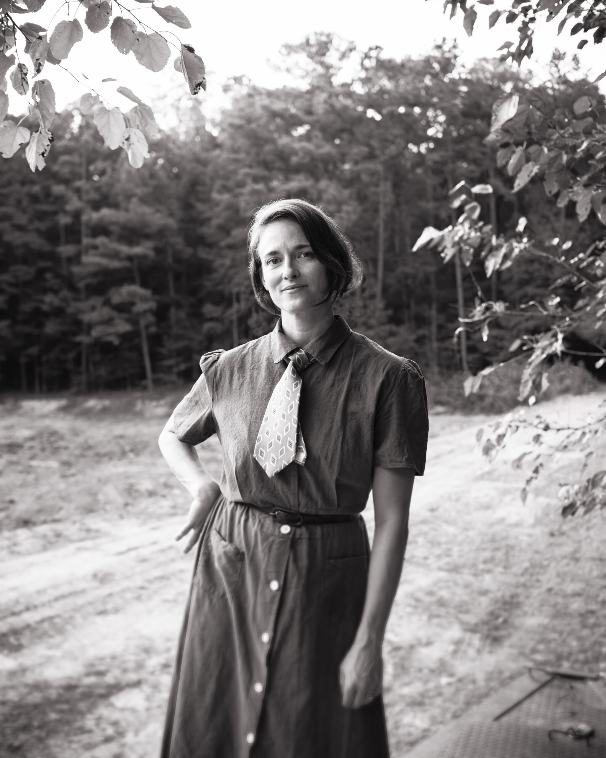 a black and white photo of a woman in a tea dress and tie standing by a tree