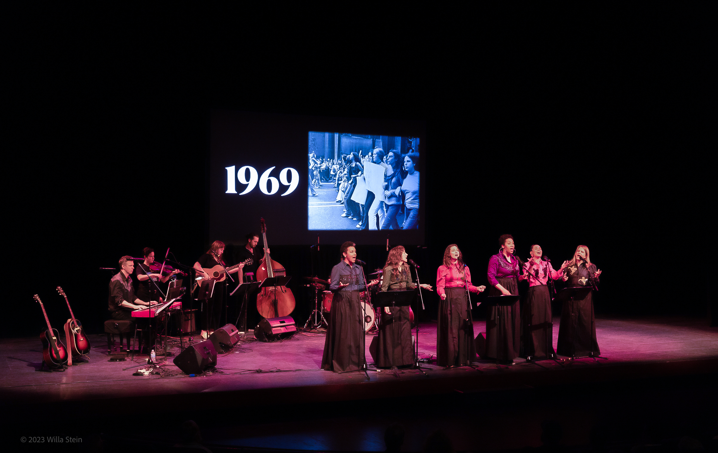 a large group musical performance featuring six female vocalists and band.