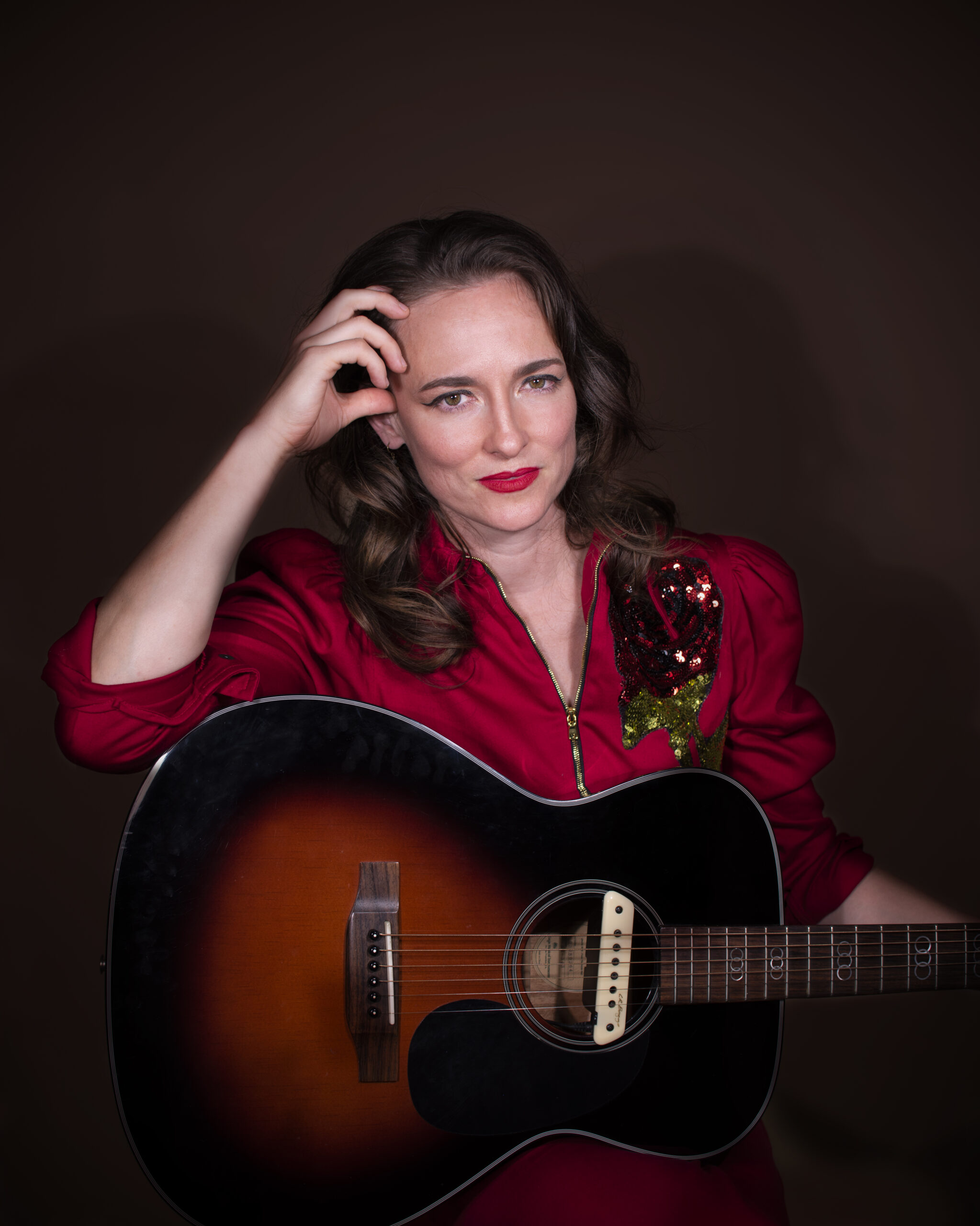 Headshot of a woman in a red blouse with dark hair and red lipstick holding a guitar