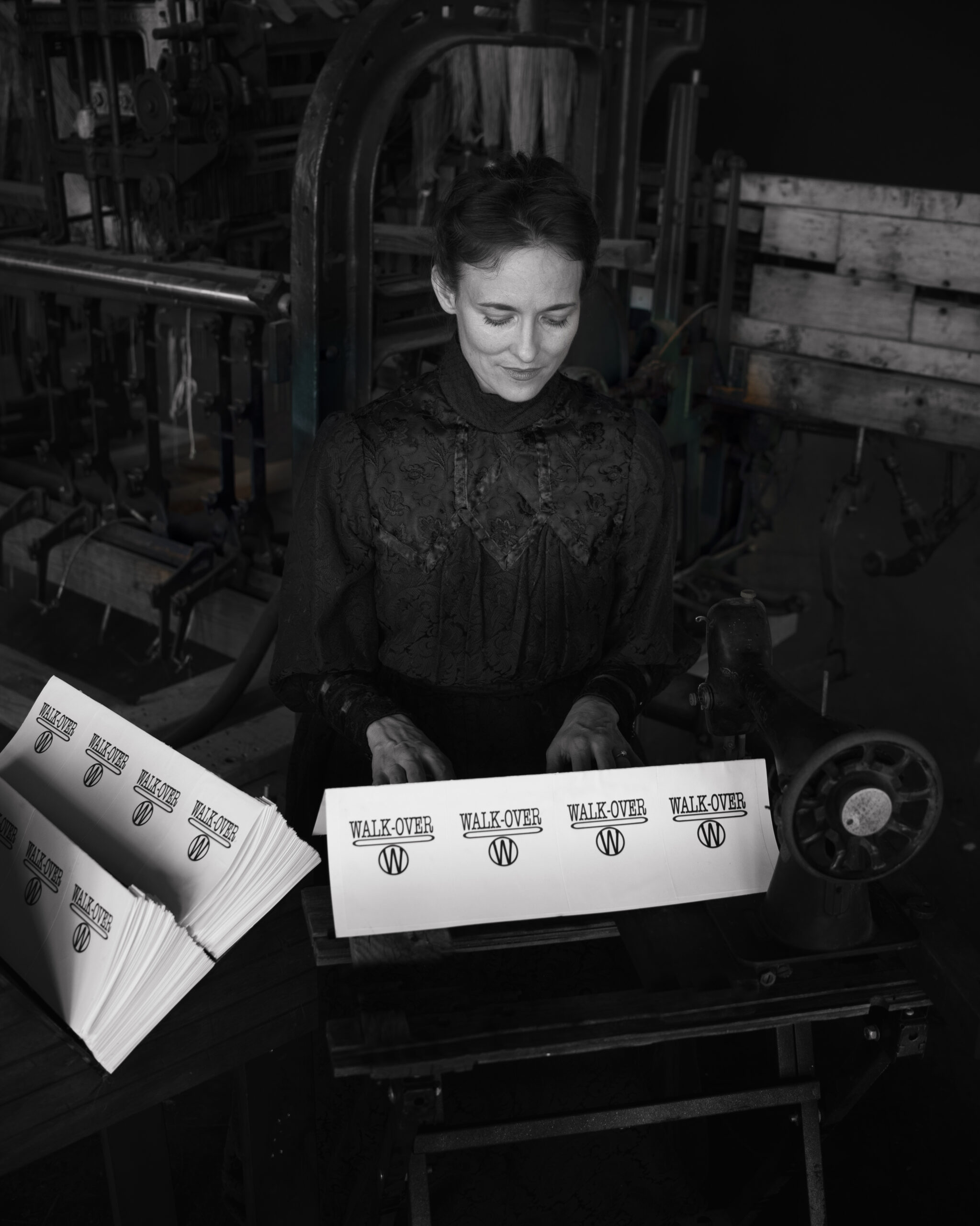 black and white photo of a woman in turn of the century attire in front of an industrial loom.
