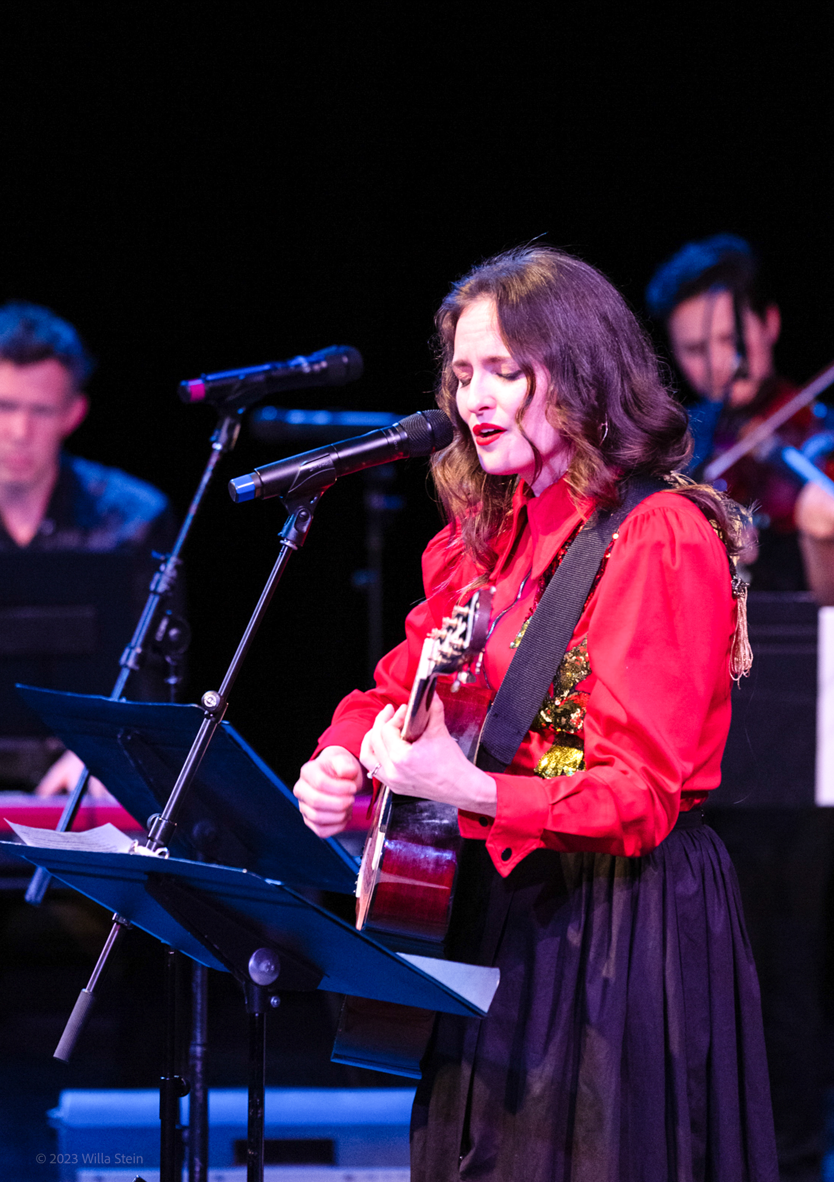 A woman in a red blouse behind a microphone singing and playing guitar live.