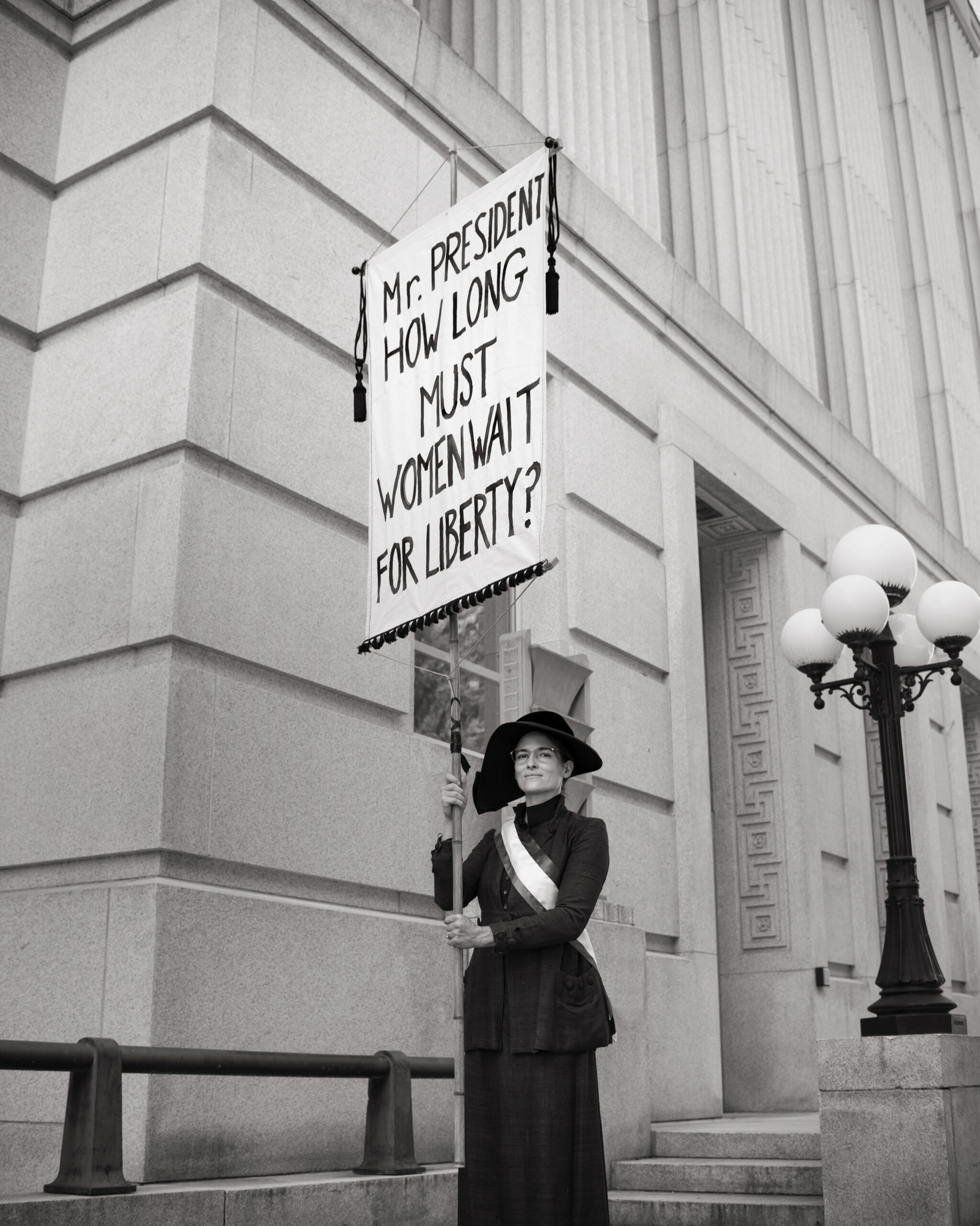 black and white photo of a suffragette holding a sign that says "mr president how long must we wait for liberty?"