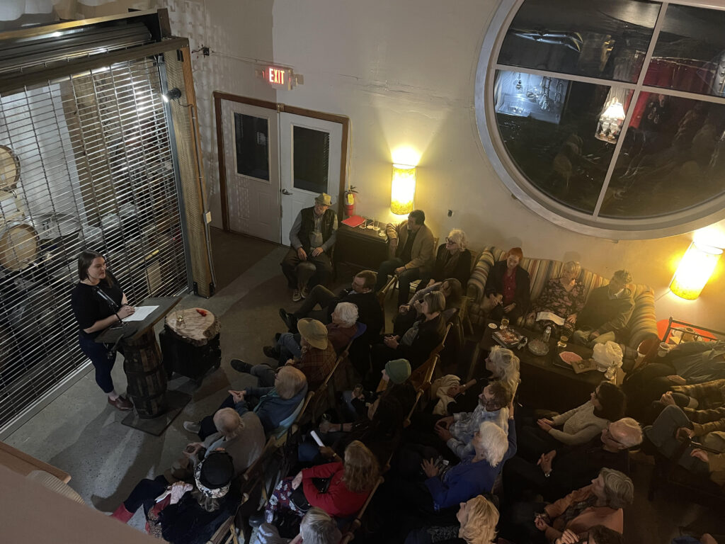 Arial photo of crowd of people listening to white woman standing behind a podium; a large round window appears in the top right