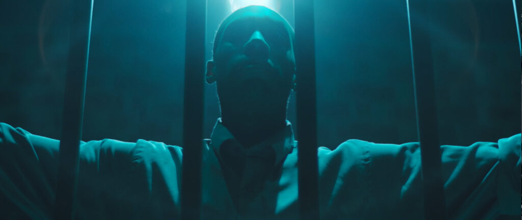 Photo of man standing behind bars with arms stretched out. Blue background light