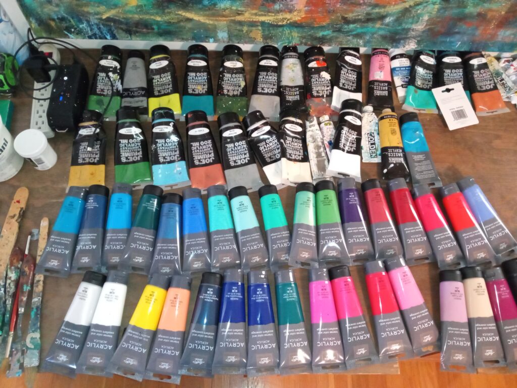 Roughly 50 fresh new tubes of acrylic paint arrayed on a wooden surface, ready for an artist to paint.