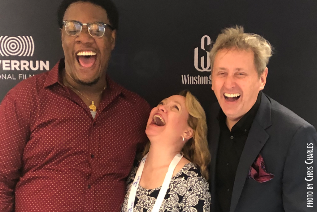 Photo of tall black man in red shirt, shorter white woman with blond hair, blonde white man in gray suit, all laughing heartily
