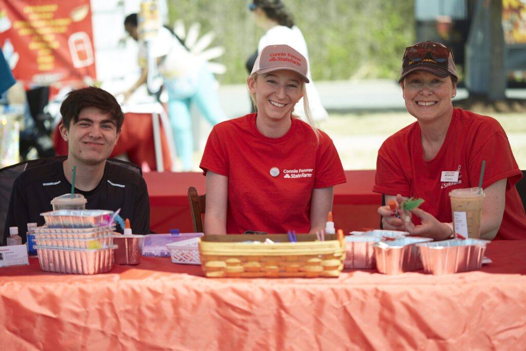Three people sitting at a table with an orange table cloth. The man on the right is wearing a black shirt and the two women on the right are wearing red shirts with the Connie Fenner State Farm logo on them.