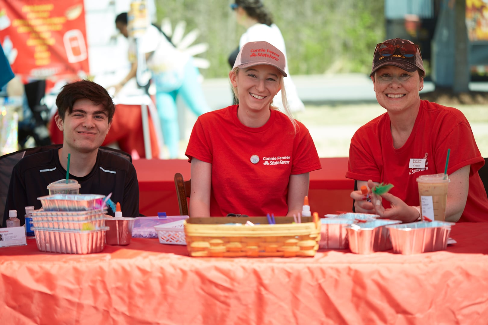 Three people sitting at a table with an orange table cloth. The man on the right is wearing a black shirt and the two women on the right are wearing red shirts with the Connie Fenner State Farm logo on them.