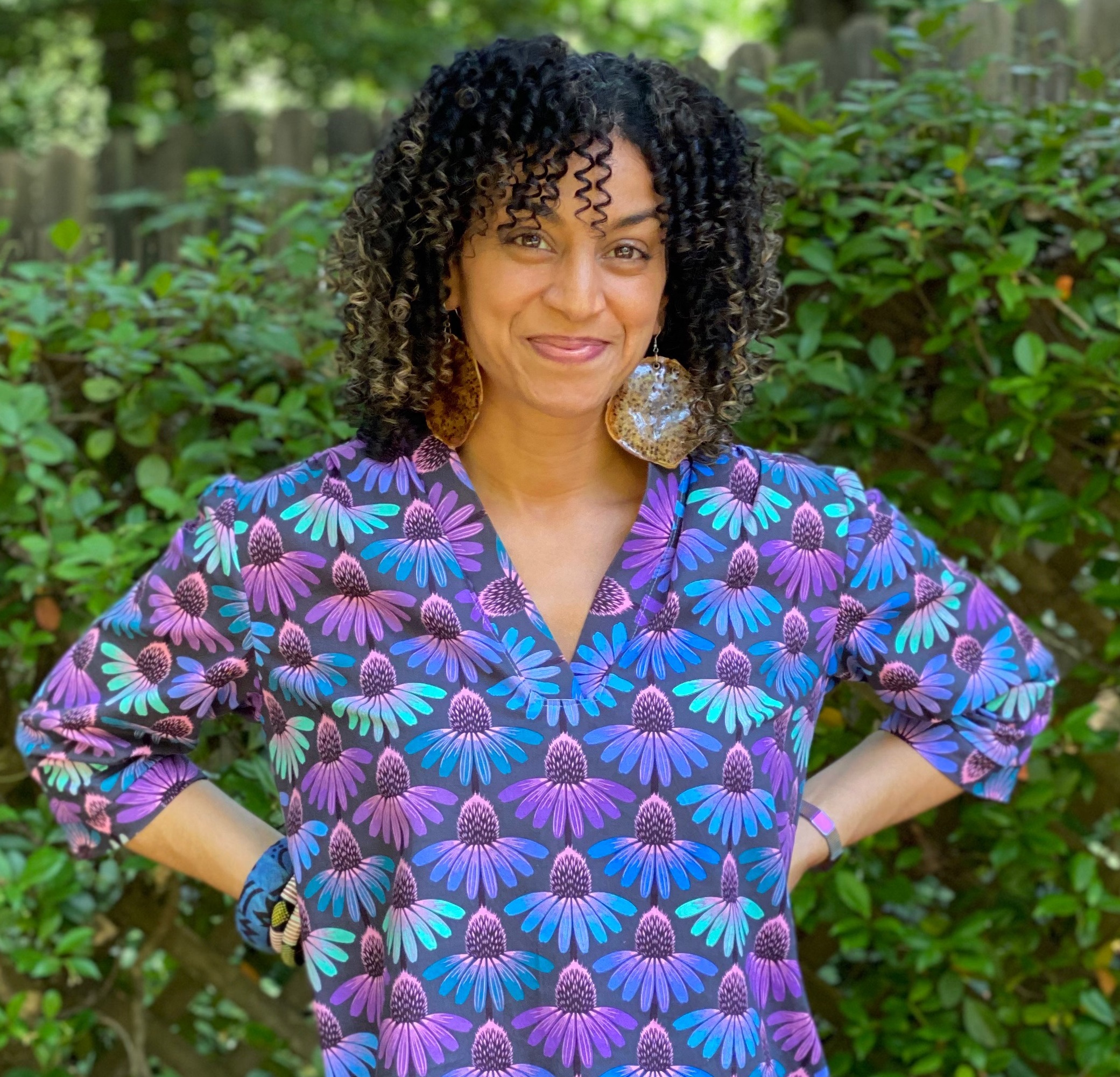 Smiling woman in a blue, purple, and pink patterned top. She is outside and her hands are on her hips. She seems happy.